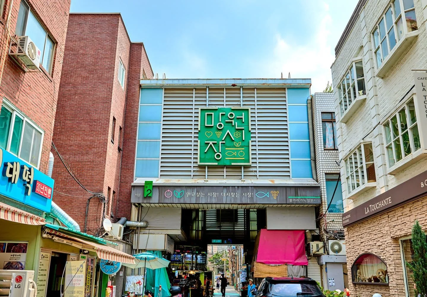 망원시장 입구 모습, 초록색 건물 지붕 위에 흰색으로 '망원시장' 간판이 있고, 그 밑으로 harmony & pleasant, mangwon 이라고 적혀있고, 그 밑으로는 서울 마포구 민방위전자고지수령&사이버교육안내 스크린이 걸려있고, 그 옆에는 망원시장이라고 예쁜 글씨로 적혀있다. 망원시장 양옆으로는 건물이 있다.